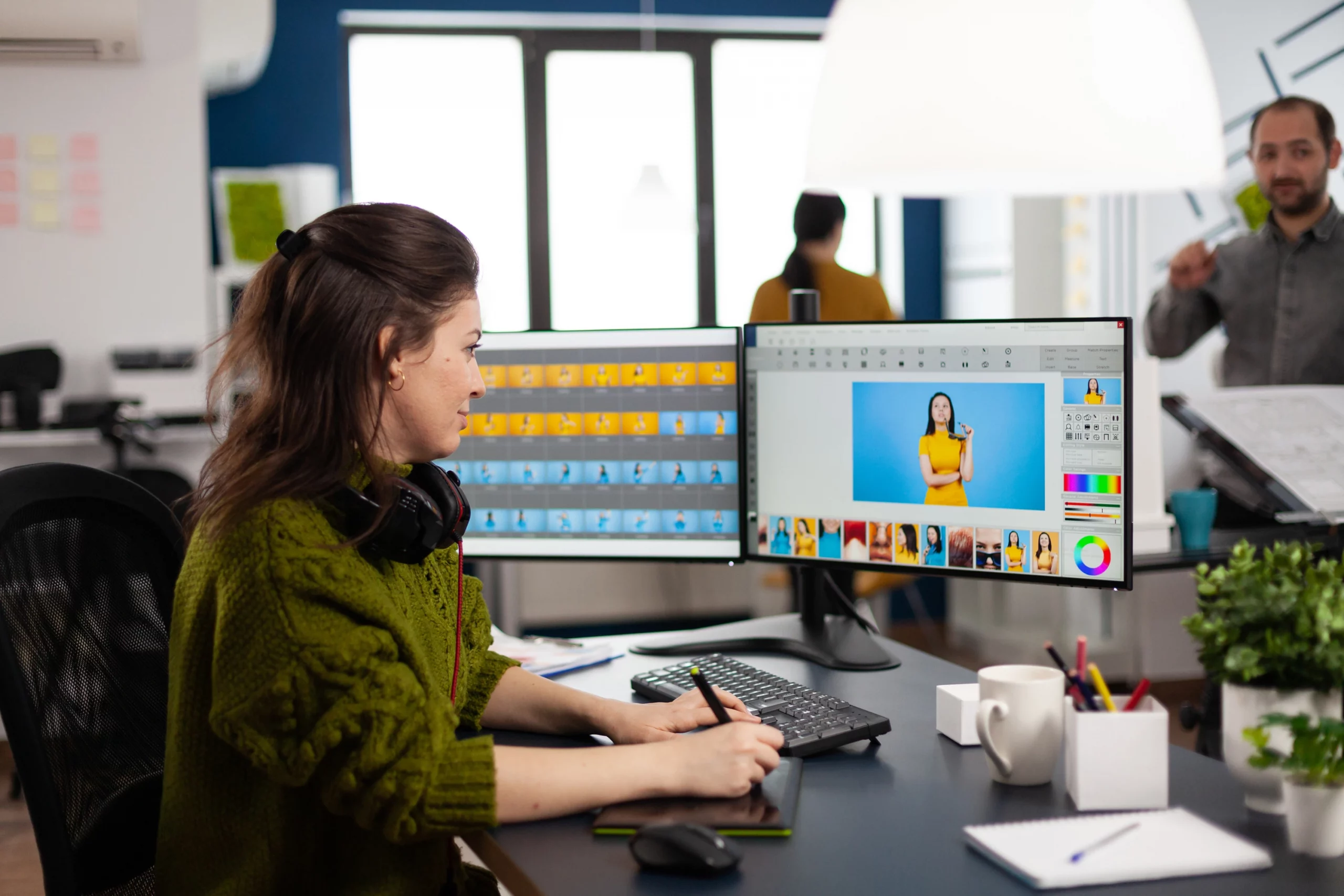 Bildbearbeiterin sitzt an einem Arbeitsplatz mit zwei großen Monitoren und optimiert ein Porträtfoto mithilfe professioneller Retusche-Software; im Hintergrund arbeiten weitere Teammitglieder, was den Prozess hochwertiger Reproduktionen und präziser Bildbearbeitung zeigt.