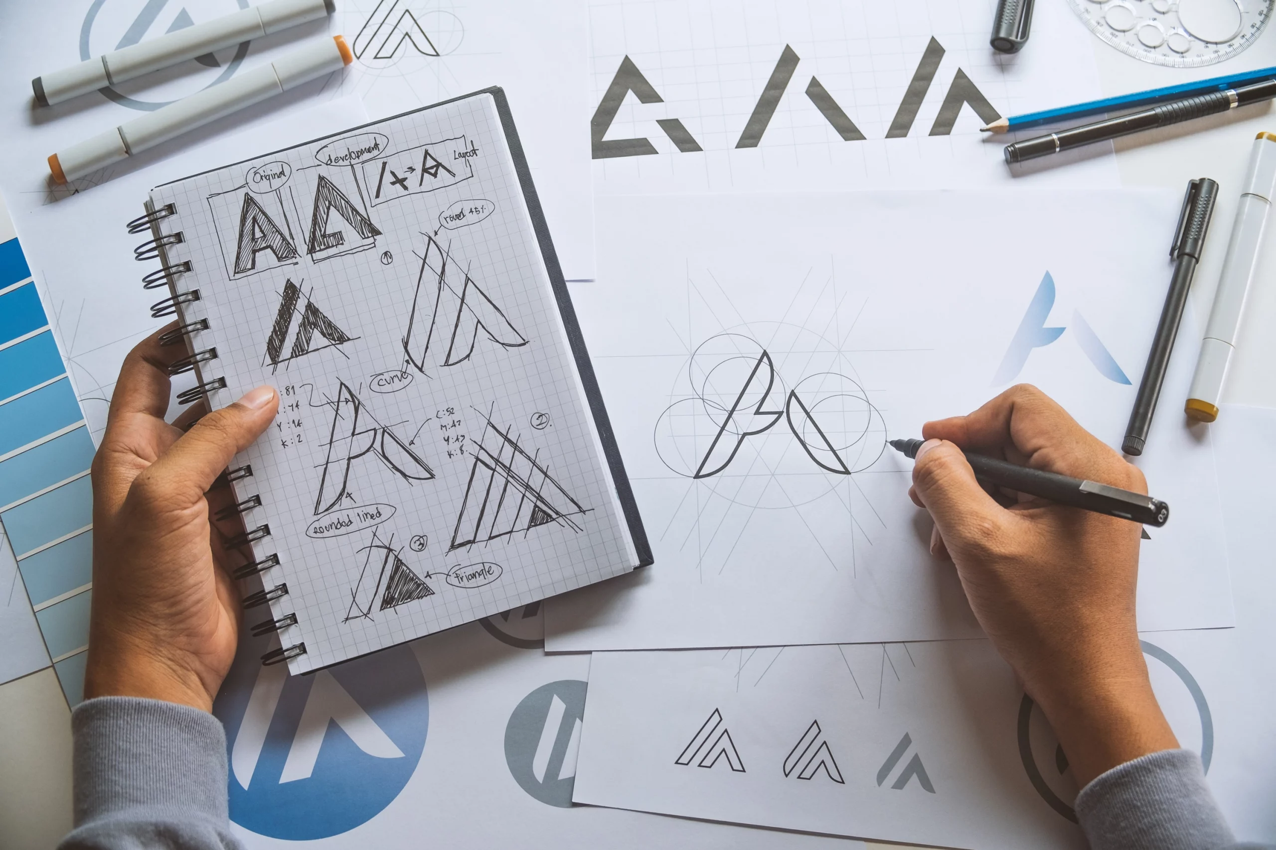 Designer skizziert verschiedene Logo-Entwürfe und geometrische Formen auf Papier, daneben ein Notizbuch mit weiteren Logos und Gestaltungsideen; die Szene zeigt den kreativen Prozess der Logo- und Icon-Entwicklung sowie die Gestaltung einer einzigartigen und konsistenten Markenidentität.