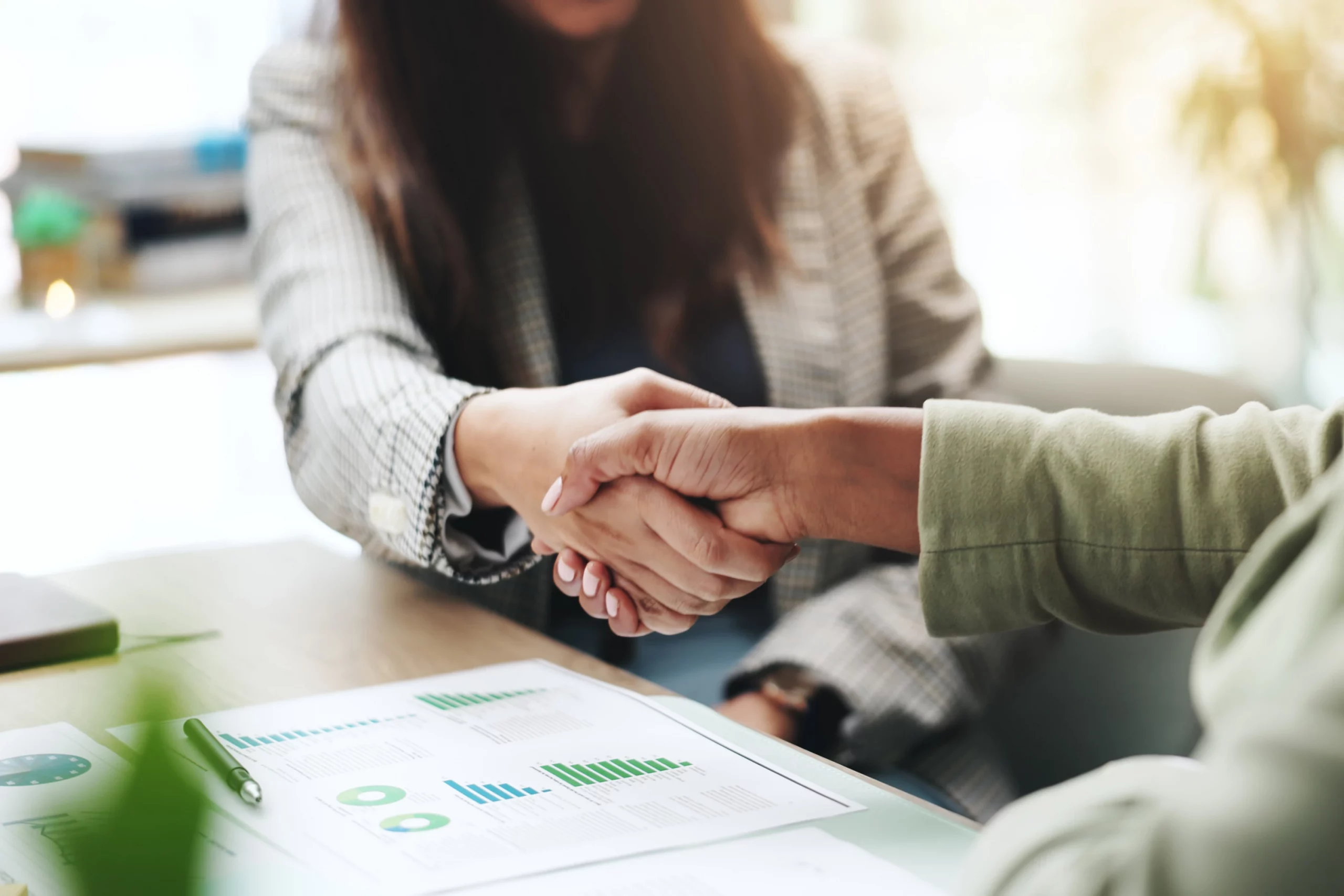 Zwei Personen reichen sich zur Begrüßung oder zum Abschluss eines erfolgreichen Gesprächs die Hand; im Vordergrund liegen Unterlagen mit Diagrammen, was eine positive Rückmeldung und den erfolgreichen Eingang einer Anfrage symbolisiert