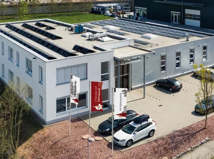 Luftaufnahme des Systemedia-Firmensitzes mit modernem Bürogebäude, Parkplatz und Firmenflaggen vor dem Eingang; die Darstellung vermittelt Professionalität und zeigt die Unternehmenszentrale.