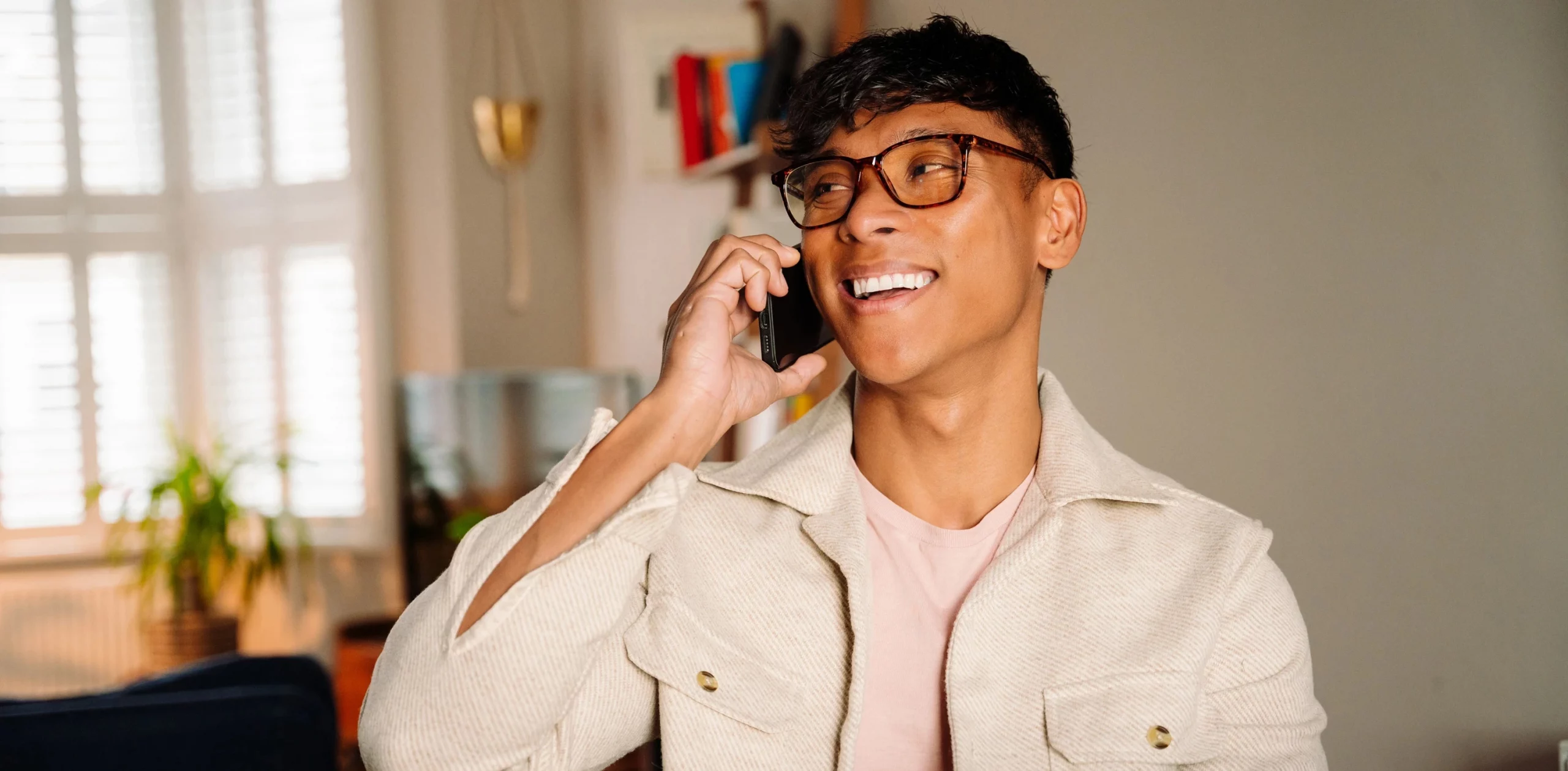 Junger Mann mit Brille sitzt in einem hellen, modern eingerichteten Raum und telefoniert lächelnd; die Szene vermittelt eine freundliche Kontaktaufnahme und passt als visuelles Element für einen ‚Kontakt aufnehmen‘-Call-to-Action.