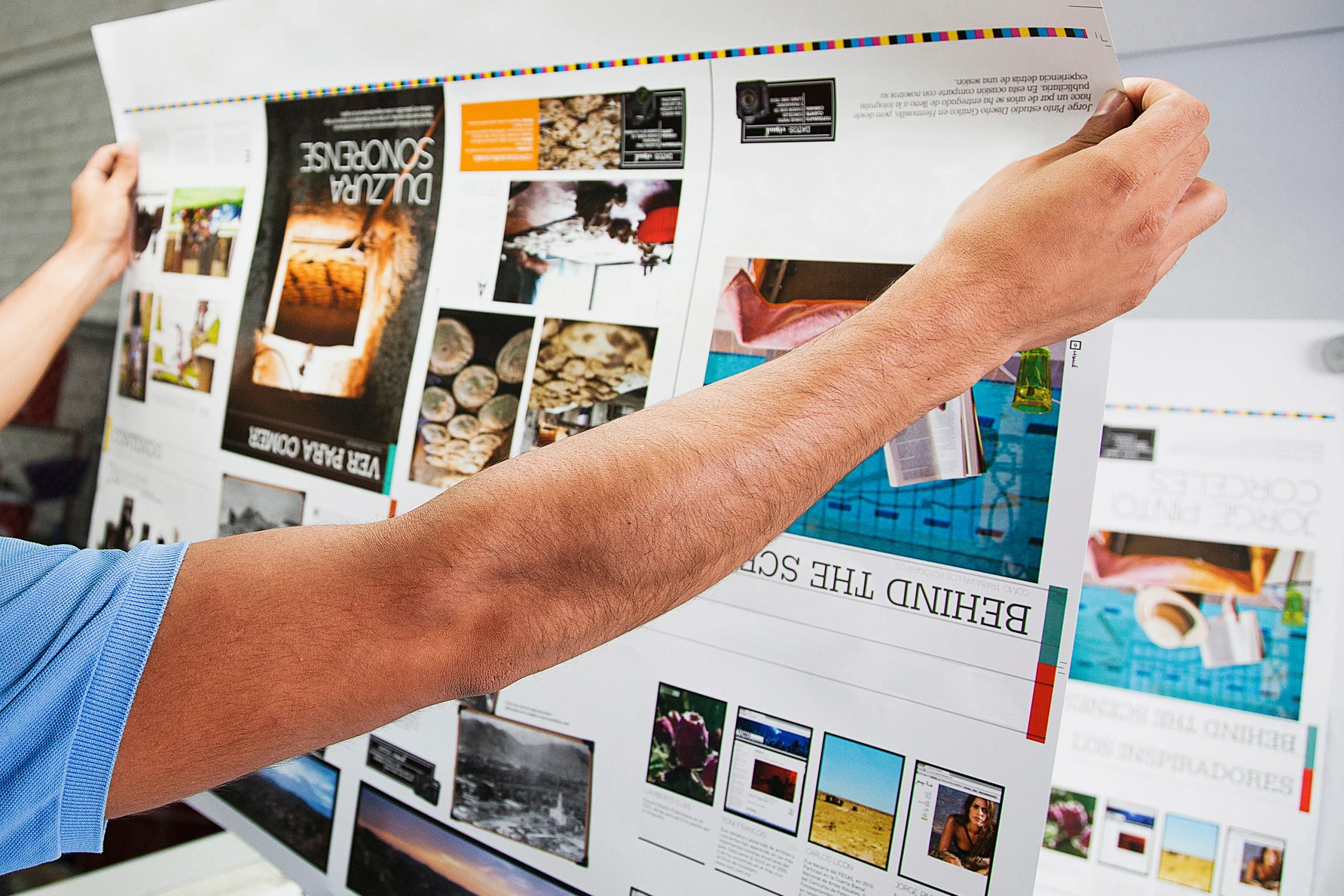 Eine Person hält einen großformatigen Druckbogen mit farbintensiven Layouts und Bildern in den Händen; die Szene zeigt den Qualitätscheck während der professionellen Print-Produktion, bei dem Farben, Details und Druckergebnis sorgfältig geprüft werden.