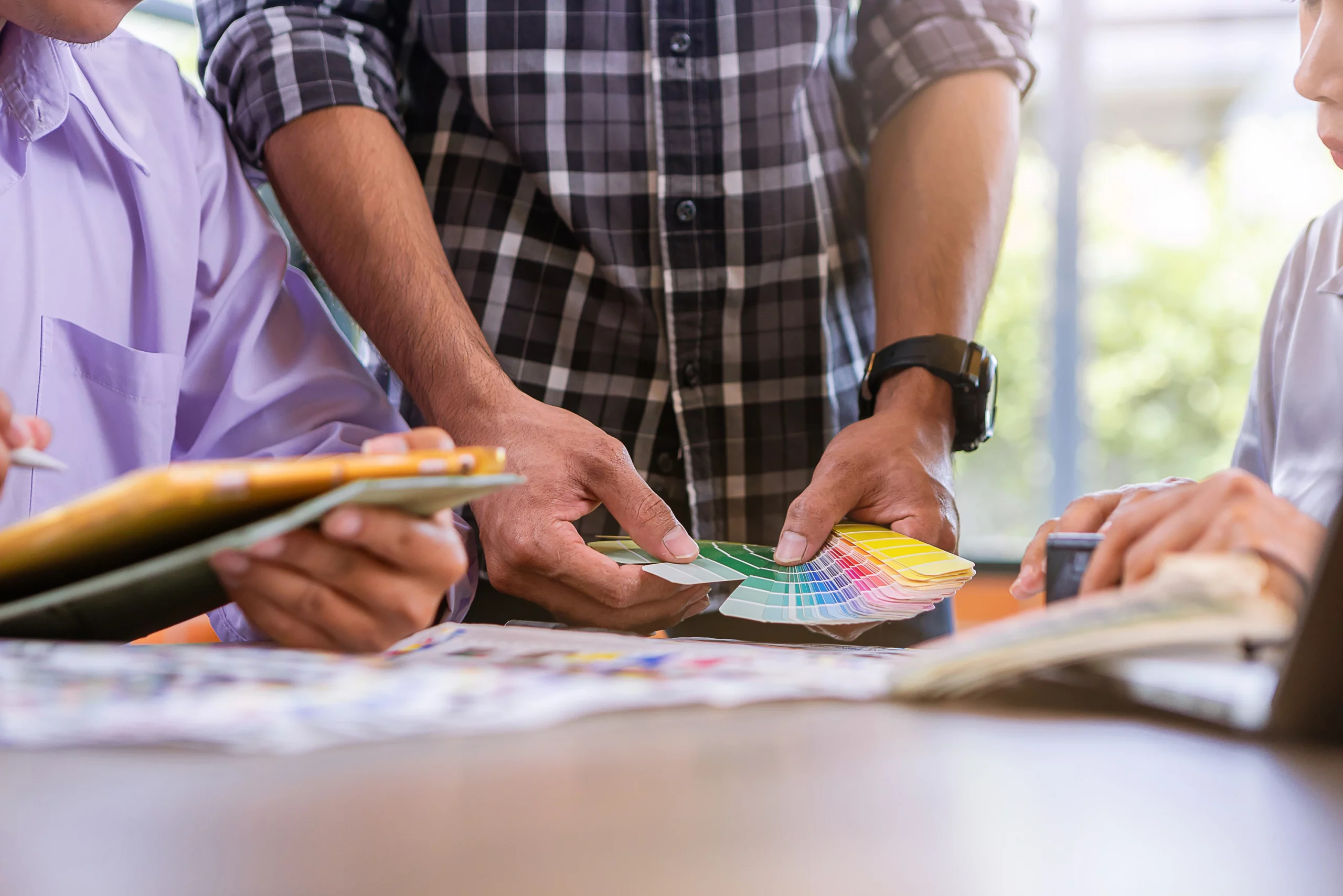 Personen prüfen gemeinsam Farbproben und Farbfächer am Tisch; der Prozess des Farbproofs wird dargestellt, bei dem Farben, Materialien und Layouts vor dem Druck abgestimmt werden, um farbverbindliche und konsistente Druckergebnisse sicherzustellen.