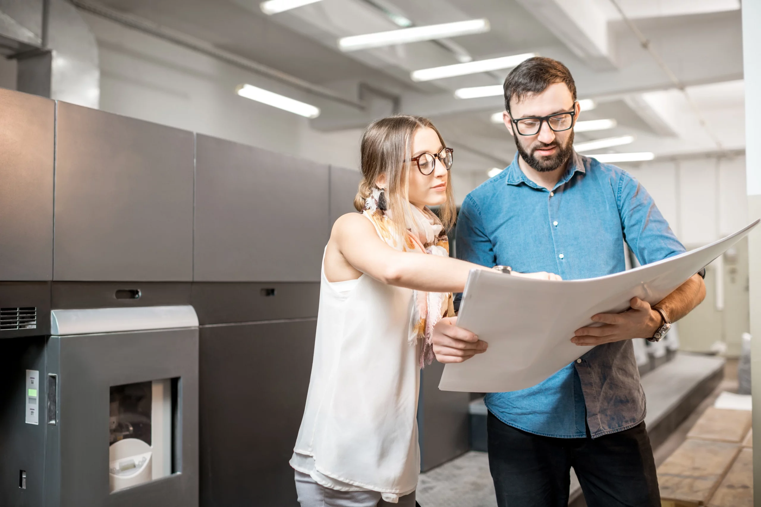 Zwei Mitarbeitende stehen in einer modernen Druckerei vor einer großen Offsetdruckmaschine und prüfen gemeinsam einen frisch gedruckten Bogen; die Szene symbolisiert präzise Druckqualität, professionelle Produktionsprozesse und hochwertige Ergebnisse im Offsetdruck